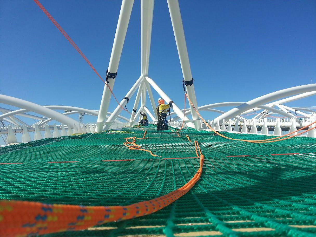 Rope access technician in hi-vis and harness on green safety netting beneath white arched steel roof trusses, under a clear blue sky