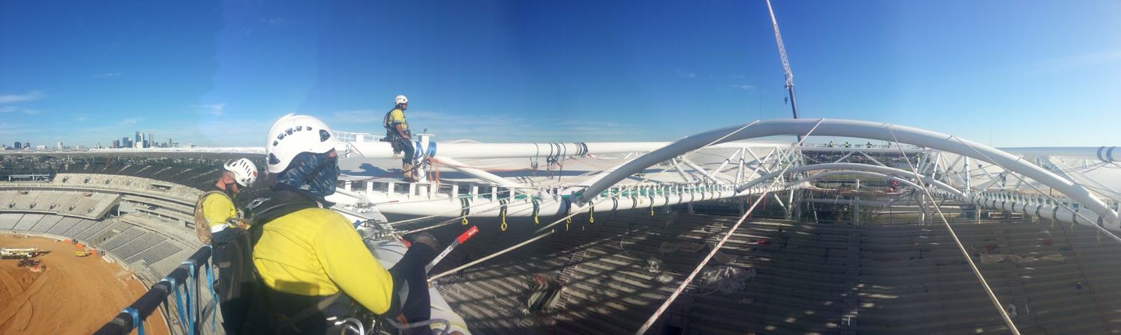 Panorama from the stadium roof: a technician in hi-vis and IRATA gear in the foreground with arched roof trusses stretching across the skyline
