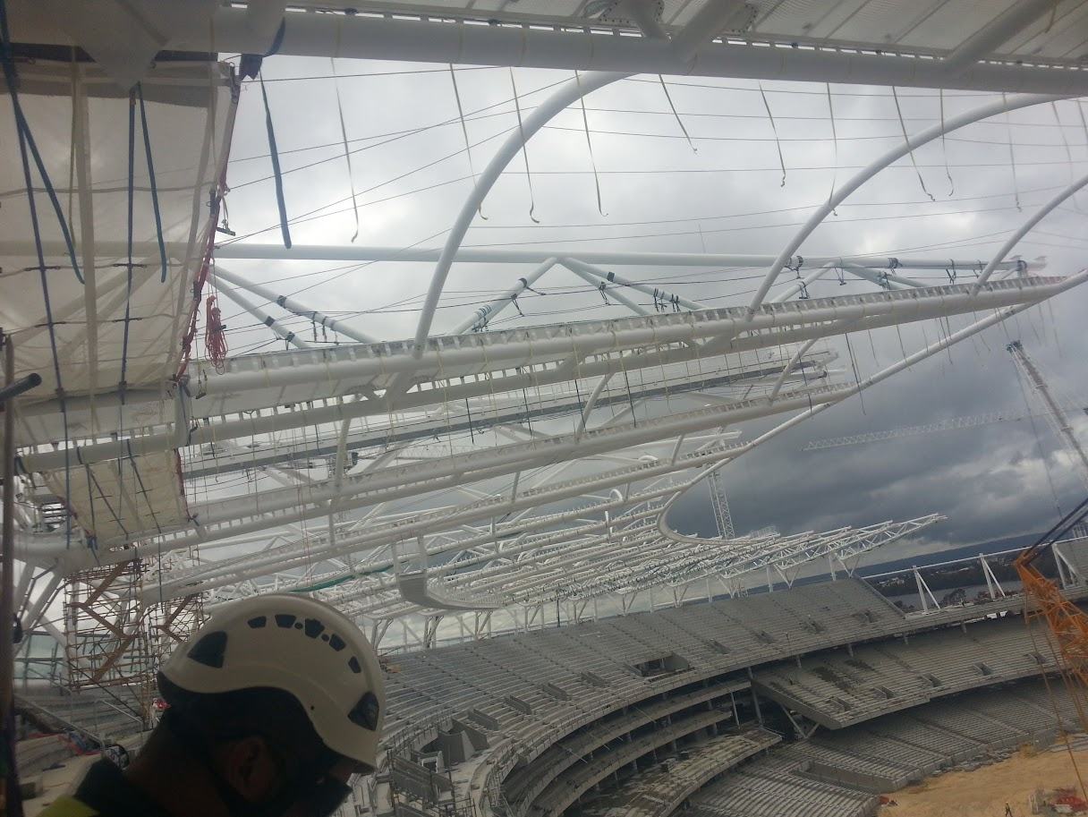 View from beneath the translucent roof membrane, looking across the steel truss framework, with a technician's helmet in the foreground