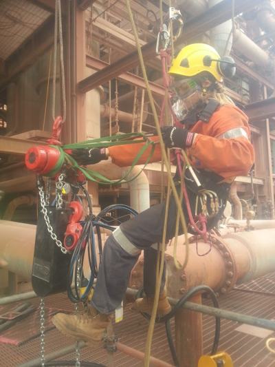Rope access technician in a respirator and orange PPE rigging a chain block on large pipework