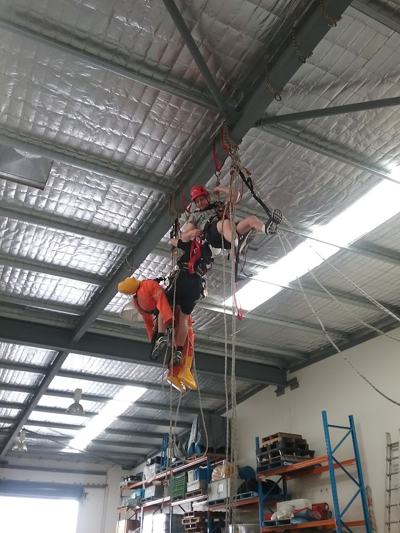 Rope rescue training inside a warehouse: technicians raising a casualty in orange PPE on rope under the roof trusses
