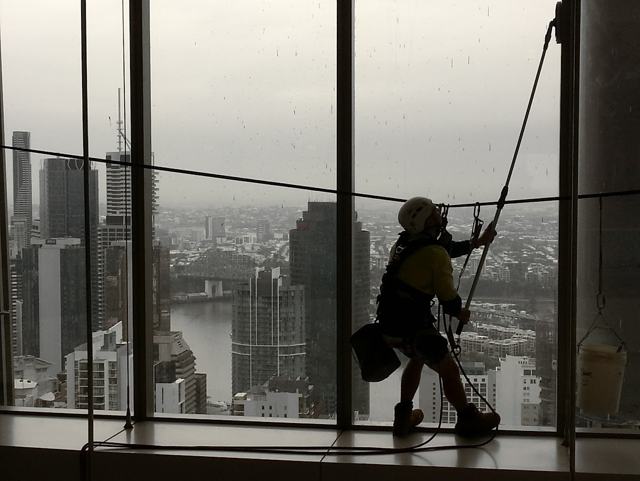 Silhouette of a rope access technician hanging at a high-rise window, with a river city skyline behind