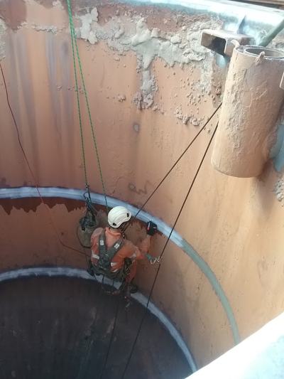 Rope access technician in orange PPE suspended inside a large rust-coloured tank, carrying out inspection and NDT work on the wall