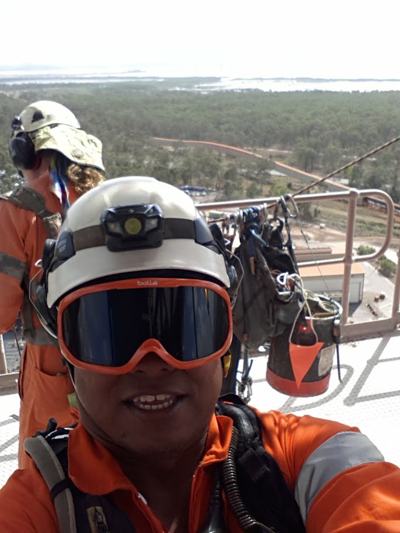 Dennis Yates at height in a white helmet and orange goggles, with a second technician behind, overlooking an industrial site